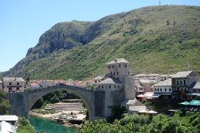 Bild von steinerner und alten Brücke in einem istorischen Ort. Dahinter eine wunderschöne Landschaft mit hohen Bergen und Wiesen.